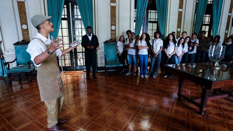 Ator caracterizado como artista histórico dirige-se aos estudantes no Salão Nobre, aproximando a plateia das vivências e do contexto social das personagens representadas.