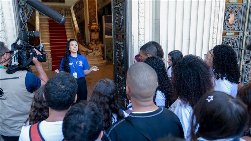Guia conduz grupo de visitantes na entrada do Palácio Tiradentes, contextualizando o roteiro e apontando como a atividade integra educação, cultura e memória.